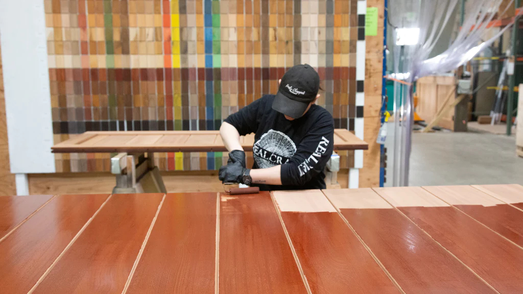 A craftswoman applies a coat of oil stain to a door with a roller.